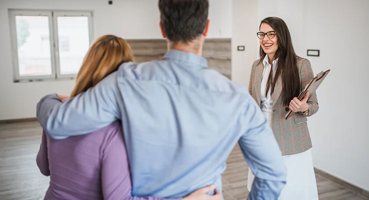The back of a couple, who have one arm around each other. A female letting agent stands opposite, facing them, smiling and holding a clipboard in one hand.
