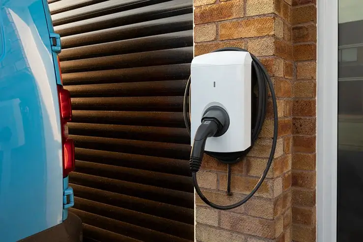 A blue electric van is parked next to a home electric vehicle charger mounted on a house wall