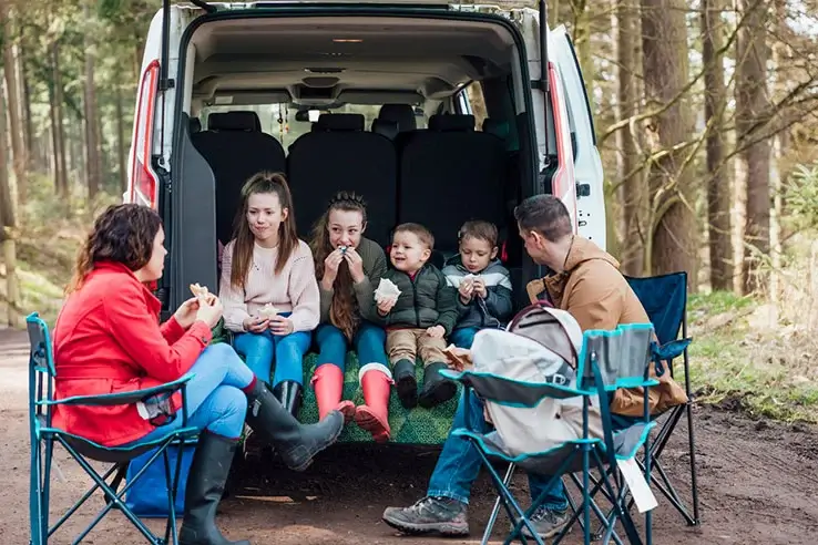Children and adults are parked in a forest, eating sandwiches around the open back doors of a van.