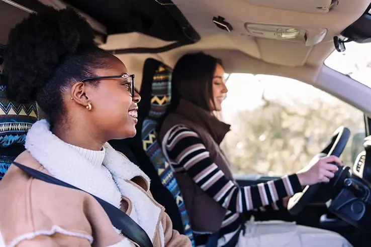 Two women are sitting in the front seats of a van, both are smiling. One is driving while the other is in the passenger seat.