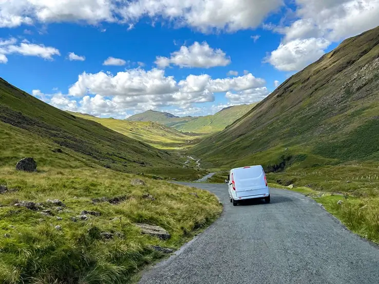 A white van is driving along a country lane that cuts through the rolling hillside.