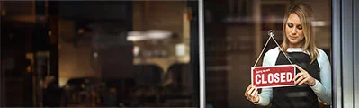 a woman wearing a black apron placing a sign on a window that reads: 'sorry we are closed'.
