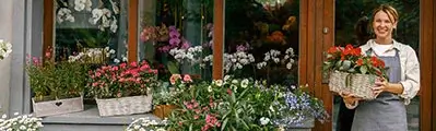 a florist standing in front of her shop holding a basket filled with flowers.