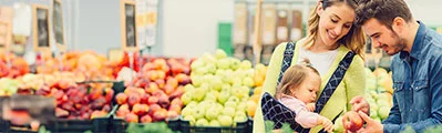 young parents with their baby daughter shopping in the vegetable aisle of a supermarket. Mother is carrying their little girl in a baby carrier.