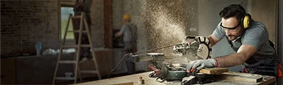 a carpenter wearing safety glasses and ear defenders is cutting wood with an electric saw on a construction site